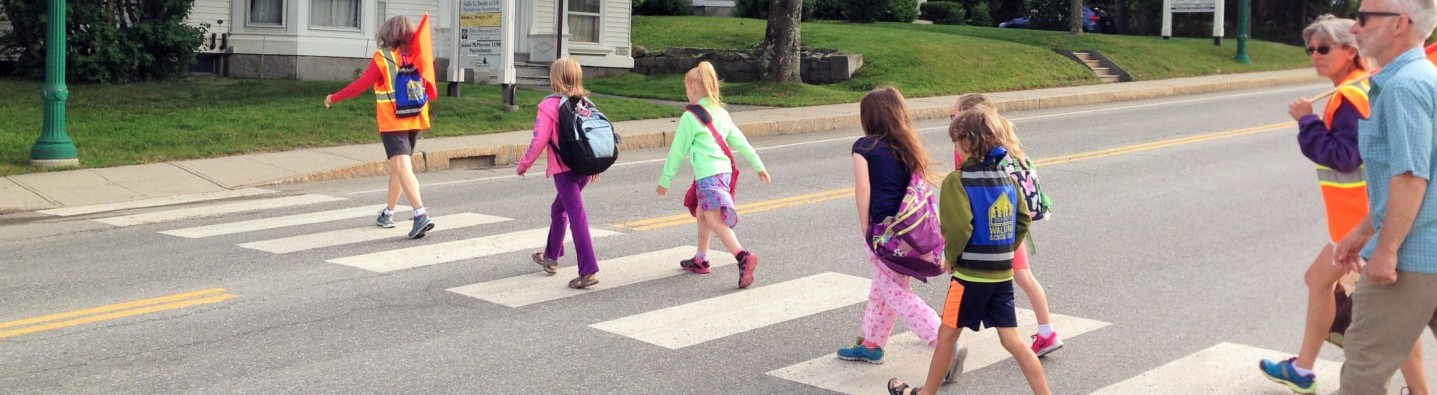 This is an image of elementary aged students walking on a crosswalk with trusted adults and a crossing guard present.